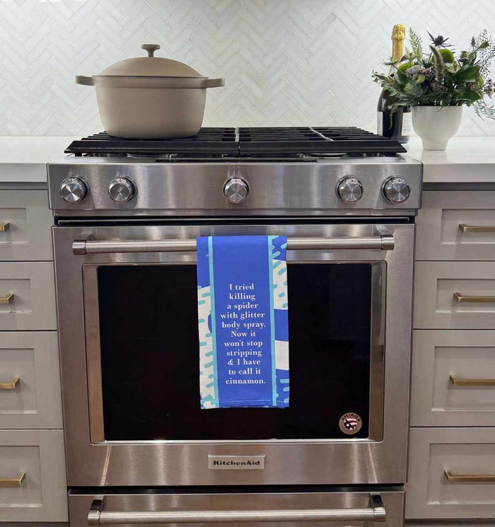 A Drinks On Me “Glitter Body Spray” dish towel hangs from a stainless steel oven handle, adding humor to your kitchen decor—perfect as a housewarming gift. A pot sits on the stove and flowers decorate the counter.