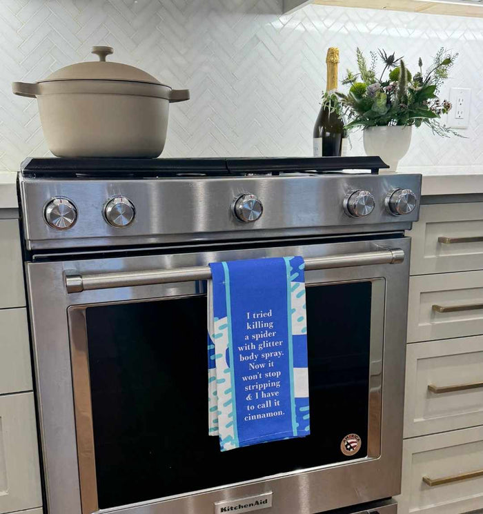 A modern stainless steel oven features the Glitter Body Spray Dish Towel by Drinks On Me, printed with a humorous kitchen quote—great as a housewarming gift. On the stove: a beige pot, bottle, and vase of flowers.