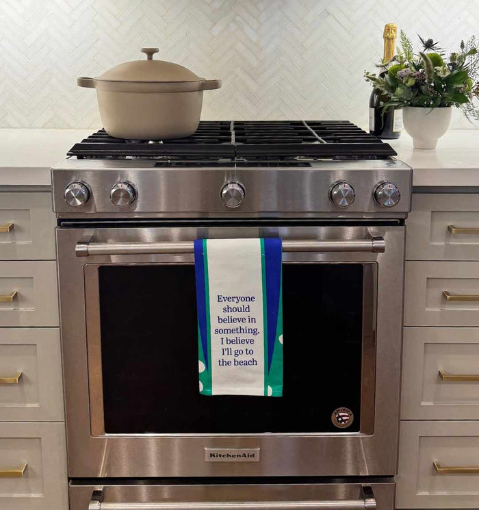 A modern stainless steel oven sports the Go to Beach Dish Towel by Drinks On Me, reading “Everyone should believe in something. I believe I’ll go to the beach”—a humorous kitchen accent and fun housewarming gift, complemented by a beige pot and plant.