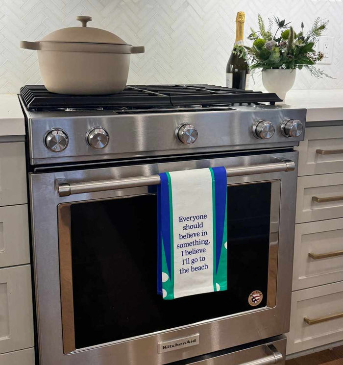 The Go to Beach Dish Towel by Drinks On Me adds kitchen humor, reading "Everyone should believe in something. I believe I’ll go to the beach." It’s a perfect housewarming gift, shown on a stainless steel oven with pot, plant, and bottle nearby.