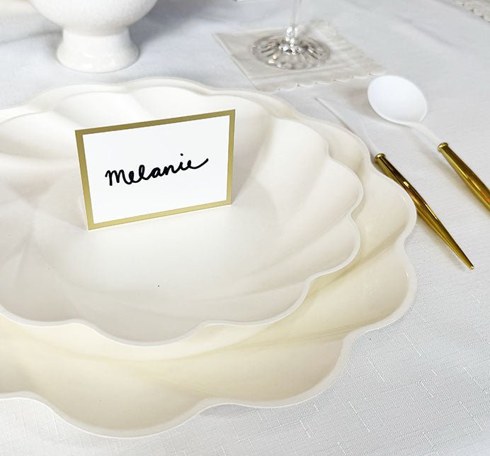 A white scalloped plate holds a Hester & Cook Gold Frame Place Card reading "Melanie" on a white tablecloth. Nearby are a white spoon, gold and white knife, fork, and wine glass on a clear coaster.