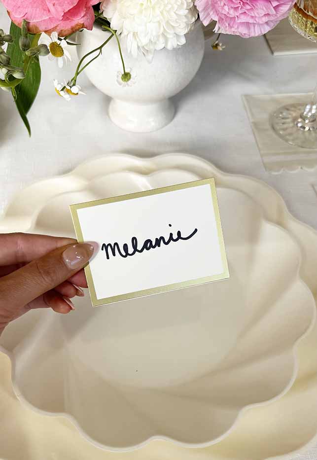 A manicured hand holds a Hester & Cook Gold Frame Place Card above a white scalloped plate, with a floral arrangement and glass in the background.