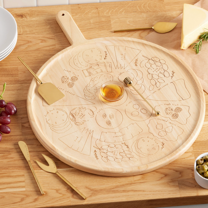A round wooden charcuterie board with labeled sections for cheeses and snacks is topped with the Maple Meg Quinn Gold Stainless Steel Honey Dipper and gold cheese utensils. Grapes, olives, and a cheese wedge sit nearby on a wooden table.