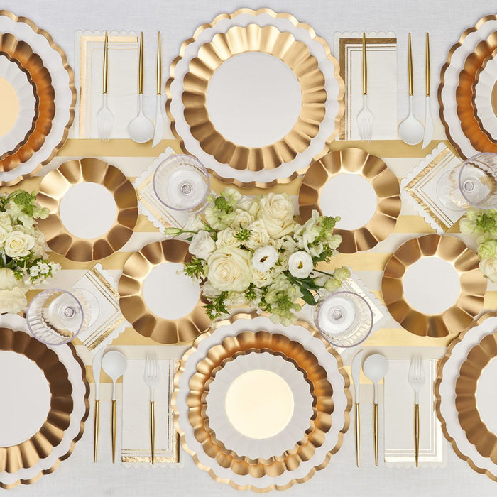 The Gold & White Table Setting by Sophistiplate LLC features gold and white scalloped designer disposable plates, gold cutlery, white napkins, and crystal glasses. Elegant white rose and greenery floral arrangements complete the table decor.