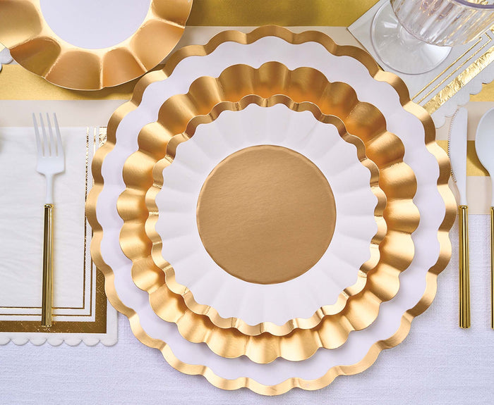 The Gold & White Table Setting by Sophistiplate LLC features designer scalloped gold and white disposable plates, gold flatware, a white napkin with a gold border, and a clear cup on a crisp white tablecloth.