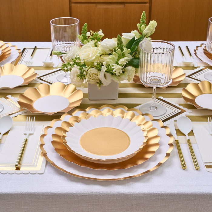 Set your dining table in style with the Gold & White Table Setting by Sophistiplate LLC, featuring designer scalloped plates, gold cutlery, white napkins with gold borders, ribbed glasses, and a white floral centerpiece on a striped runner.