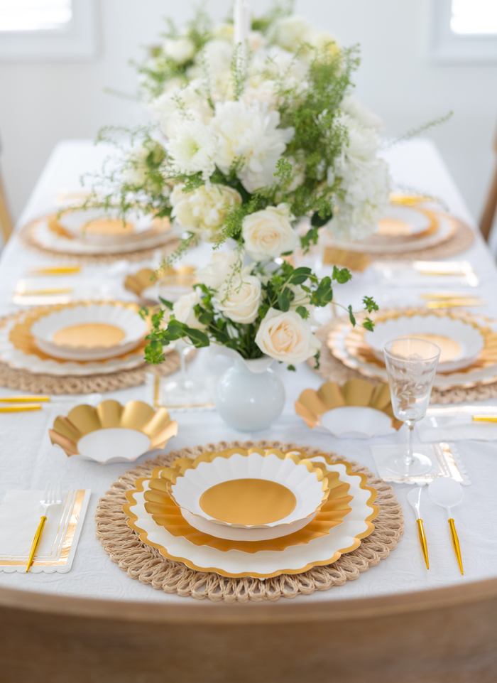 The Gold & White Table Setting by Sophistiplate LLC features designer disposable plates with gold and white scallops, gold utensils, crystal glasses, a centerpiece of white roses and greenery in a white vase, woven placemats, and a white tablecloth.