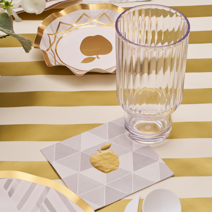 A ribbed glass sits on a gold and white striped tablecloth with gold-accented plates and a Sophistiplate Golden Apple Cocktail Napkin, featuring a golden apple print. White plastic cutlery appears at the edge, completing this elegant table setting.