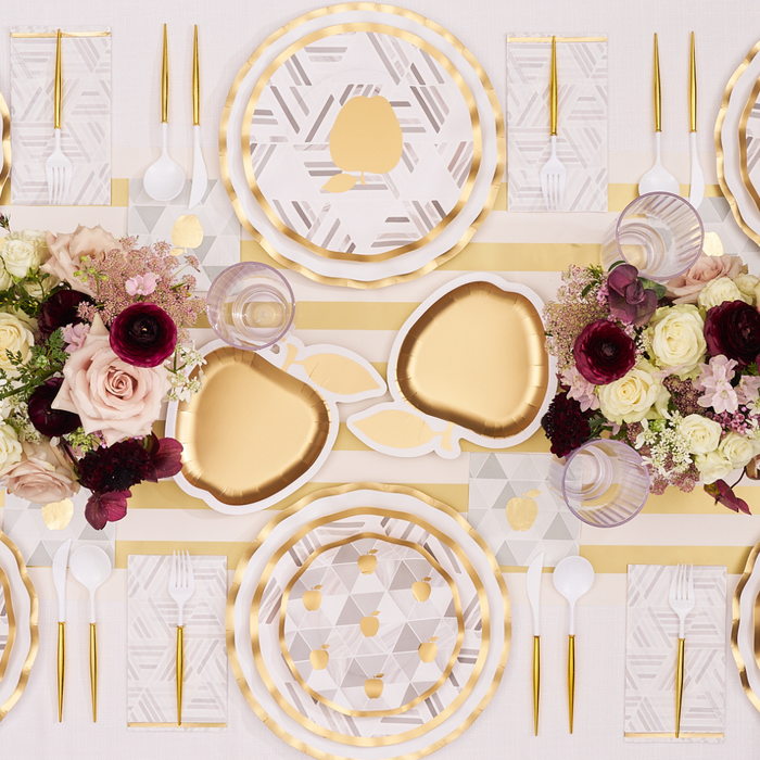 A formal Golden Apple Table Setting features gold and white plates—including Sophistiplate Golden Apple Die-Cut Salad Plates (8pk)—gold cutlery, geometric napkins, floral centerpieces with burgundy and white flowers, and gold accents on a cream tablecloth.