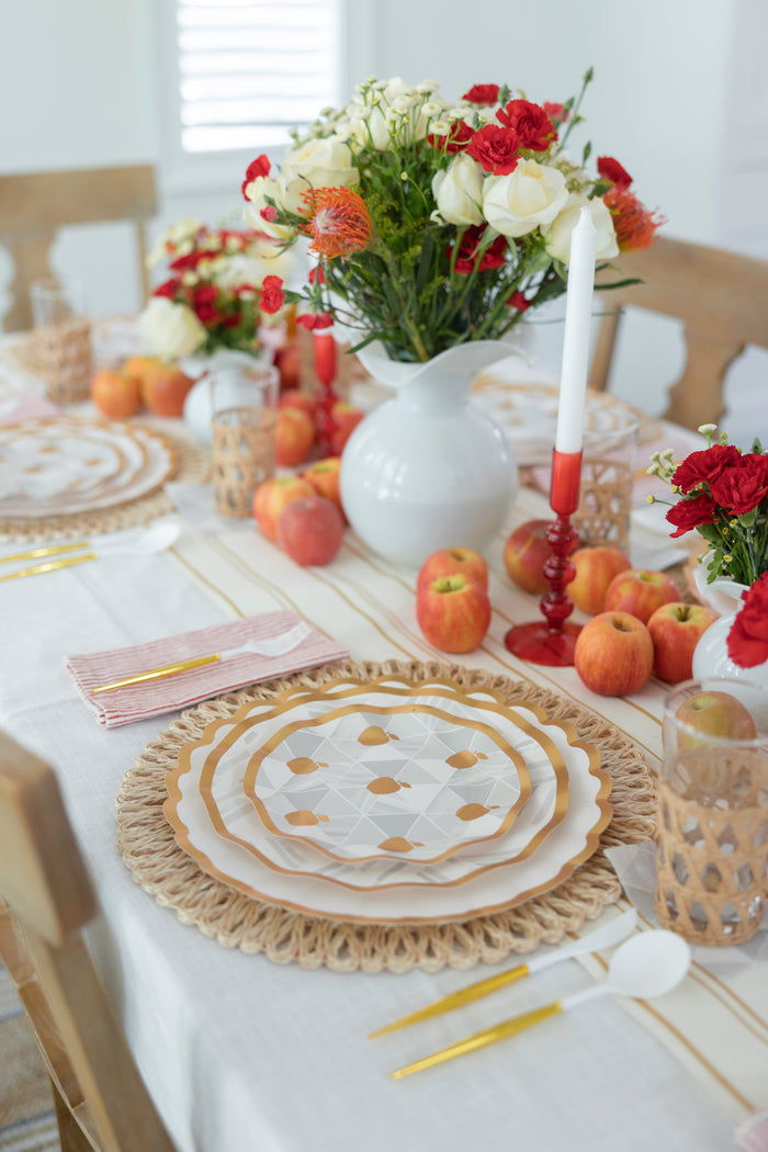 A dining table set with layered plates, gold cutlery, woven placemats, and a Hester & Cook Antique Gold Stripe Runner is decorated with vases of red and white flowers, apples, candles, and glasses for a fresh, festive touch.