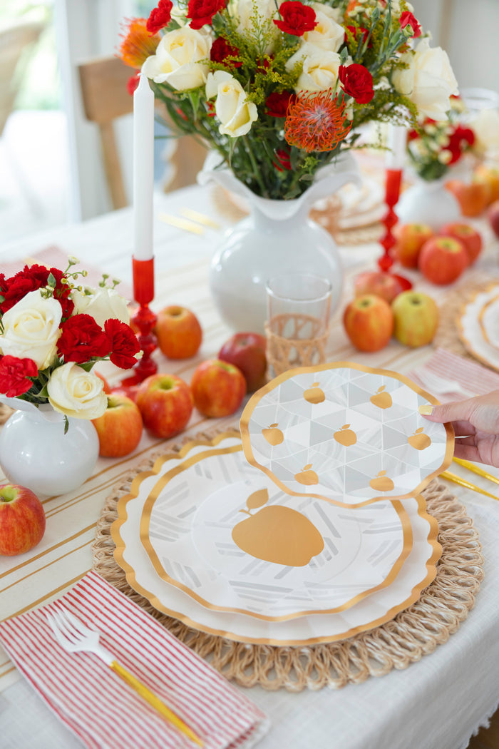 Create a stunning Rosh Hashanah table with the Golden Apple Table Setting by Sophistiplate: designer disposable plates with apple motifs, floral vases, scattered apples, white candles, red-striped napkins, gold utensils—a hand sets the plate.