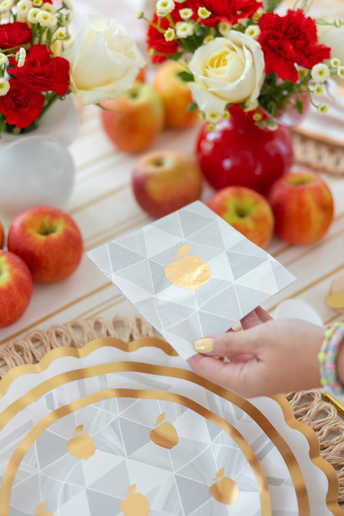 A hand holds a decorative white napkin with gold apple motifs above the Sophistiplate Golden Apple Table Setting, featuring apples, designer disposable plates, and floral vases—perfect for a festive Rosh Hashanah celebration.