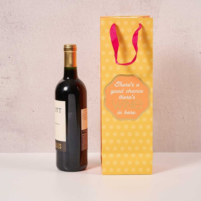 A bottle of red wine is beside the Drinks On Me Good Chance- Wine Bag, a reusable bag with yellow polka dots, pink handles, and the message “There’s a good chance there’s WINE in here,” on a white surface against a light background.