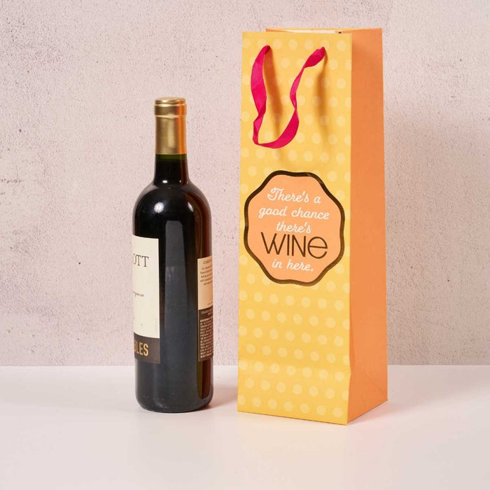 A bottle of red wine stands beside the Drinks On Me Good Chance- Wine Bag, featuring a yellow polka dot design, red handles, and the text: "There’s a good chance there’s WINE in here.
