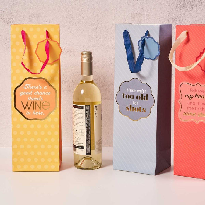 Three Drinks On Me Good Chance-Wine Bags with colorful handles stand upright, featuring funny messages, with a bottle of white wine between the first and second bag, all set against a light background.