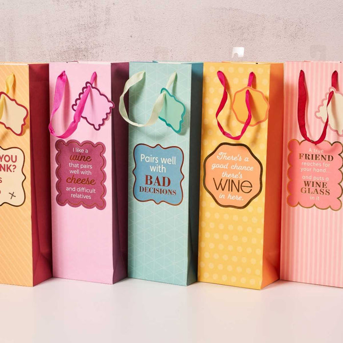 Five Drinks On Me Good Chance Wine Bags with humorous wine, cheese, and friendship phrases feature decorative tags and ribbon handles. These colorful, reusable gift bags are lined up against a light wall.