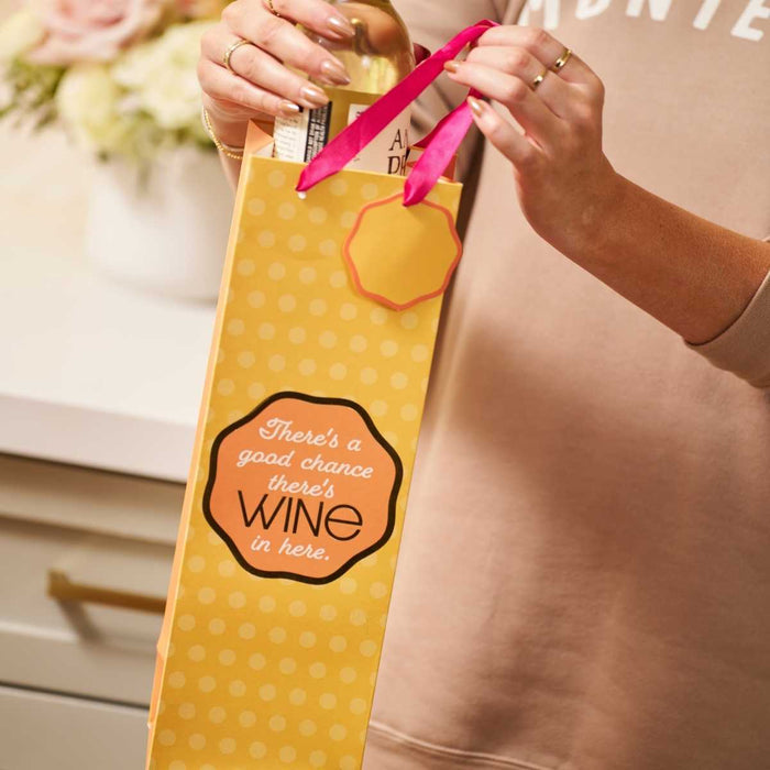 A person holds the Drinks On Me Good Chance- Wine Bag—a yellow polka-dotted gift bag with pink ribbon handles and the phrase "There's a good chance there's WINE in here"—while placing a wine bottle inside.