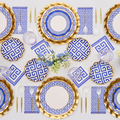 The Greek Key Table Setting by Sophistiplate LLC features blue and white Greek key plates, gold chargers, gold and clear flatware, blue patterned napkins, striped glasses, and a centerpiece with white and blue flowers.