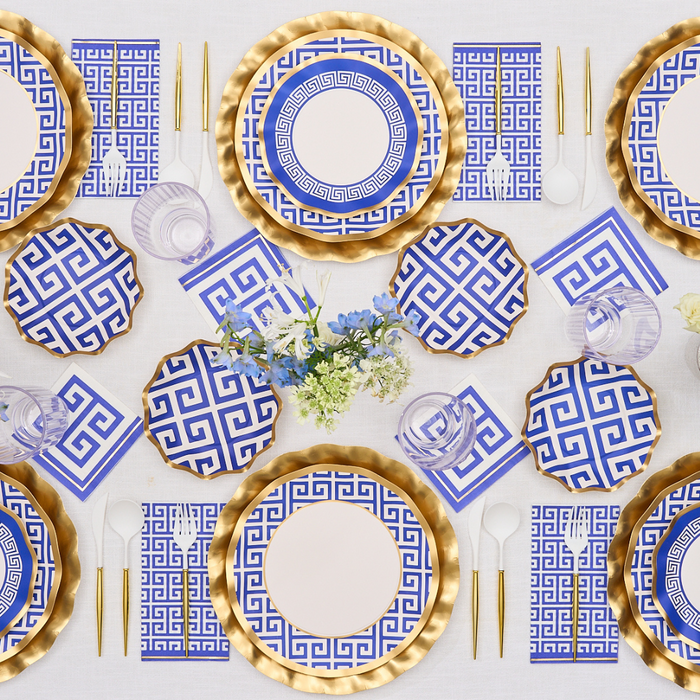The Greek Key Table Setting by Sophistiplate LLC features blue and white Greek key plates, gold chargers, gold and clear flatware, blue patterned napkins, striped glasses, and a centerpiece with white and blue flowers.