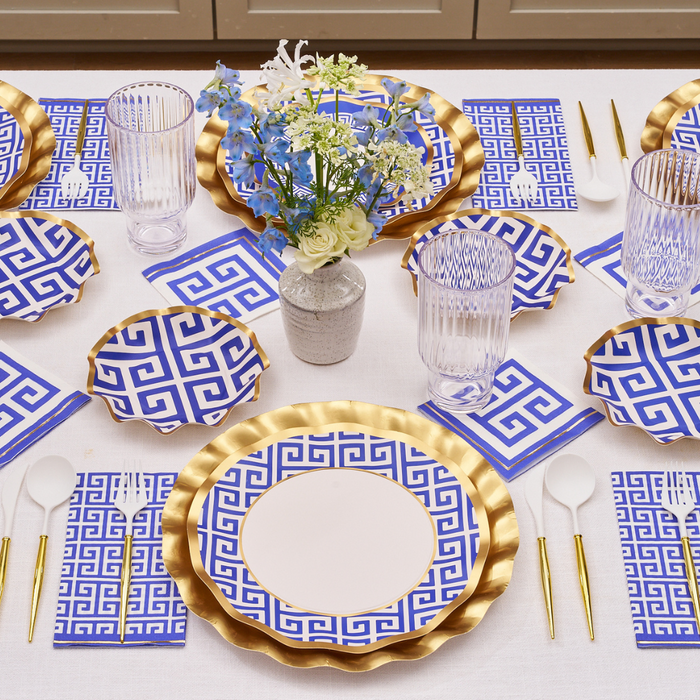 The Greek Key Table Setting by Sophistiplate LLC features gold-rimmed disposable plates, blue and white Greek key-patterned plates and napkins, clear glasses, gold cutlery, and a vase with white and blue flowers on a white tablecloth.