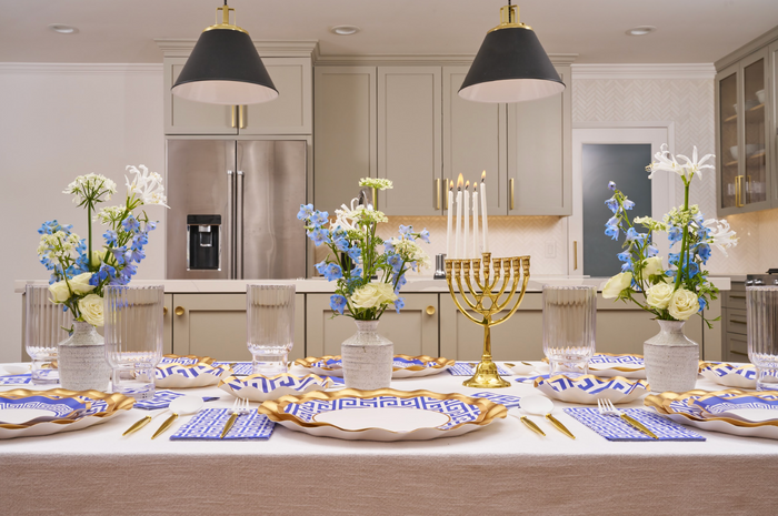 The Greek Key Table Setting by Sophistiplate LLC features blue and white floral centerpieces, gold cutlery, designer disposable plates, and a gold menorah arranged on a dining table in a modern kitchen with gray cabinets and pendant lights.