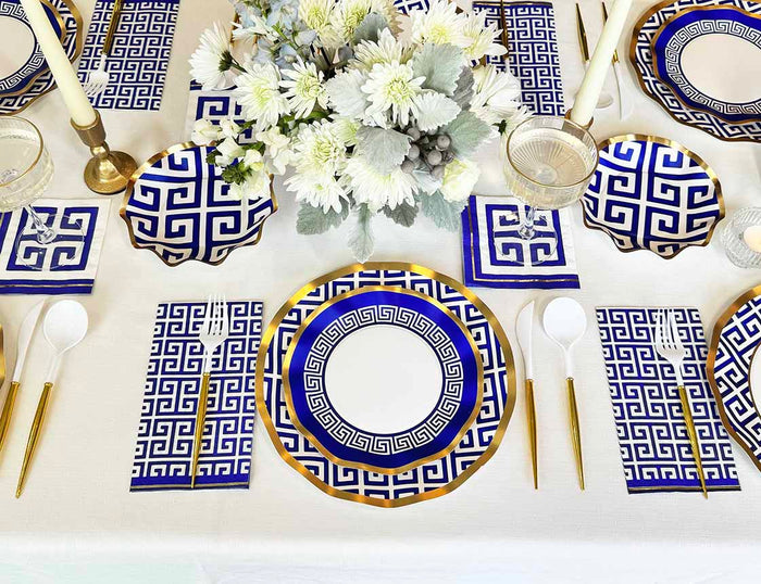 The Greek Key Table Setting by Sophistiplate LLC features blue and white designer disposable plates, bowls, napkins, and cups with gold accents. Gold flatware, brass candleholders, and a white floral centerpiece complete the elegant look.