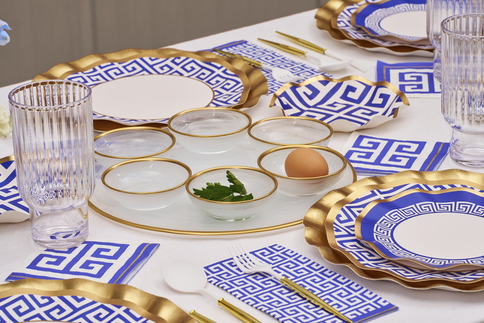 The Greek Key Table Setting by Sophistiplate LLC features blue and white designer disposable plates, napkins, a gold-edged serving tray with small bowls for herbs, water, and an egg, plus clear glasses and utensils—all neatly arranged for a formal dining look.