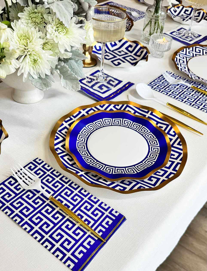 The Greek Key Table Setting by Sophistiplate LLC features designer blue and white disposable plates with gold trim, matching napkins, gold cutlery, a wine glass, and a floral centerpiece on a crisp white tablecloth.