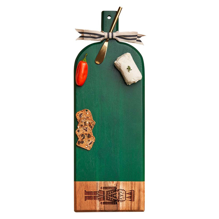 The Maple Green Acacia Bevel Board w/ Spreader (Nutcracker design, 20x7") features goat cheese, a red pepper, two crackers, a gold spreader, and a ribbon-tied handle.