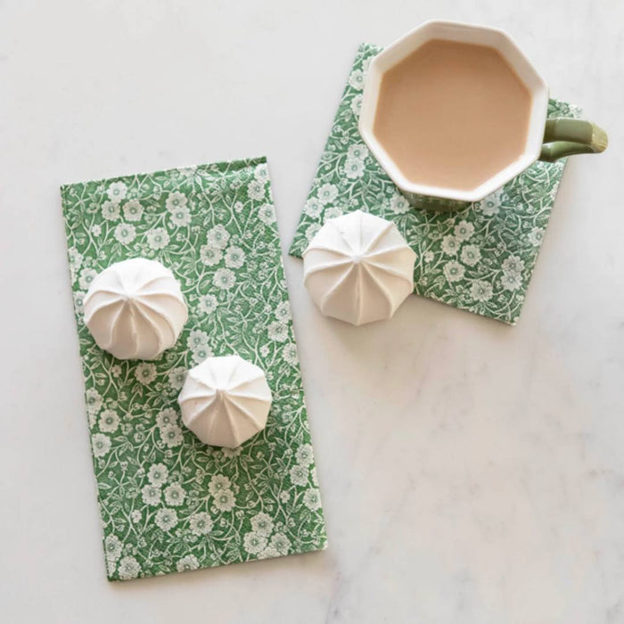 A cup of tea with milk rests on a Hester & Cook Green Calico Paper Cocktail Napkin next to three white meringue cookies, all arranged on a white marble surface.