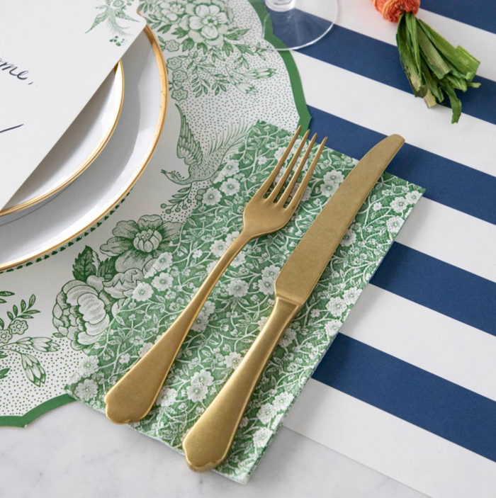 A gold fork and knife rest on Hester & Cook Green Calico Paper Guest Towels atop a blue and white striped tablecloth, next to a white plate with a gold rim and a piece of paper.