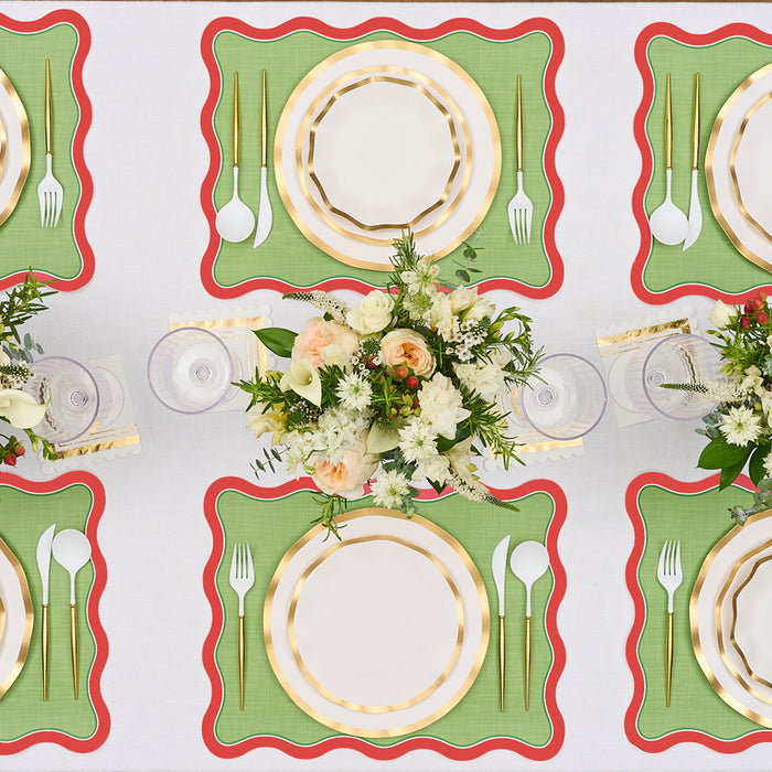 A table setting features Rosanne Beck Green & Red Scallop Edge Placemats (10pk), gold-rimmed plates, gold flatware, clear glasses, and floral centerpieces of white and peach blooms with greenery, all arranged symmetrically.