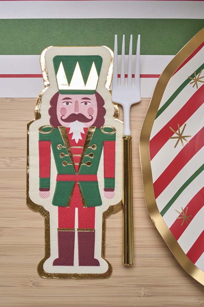 A festive table setting features Sophistiplate's Guest Towel Nutcracker Christmas paper napkins, a white fork with a gold handle, and a plate decorated with red, green, and gold stripes and stars.