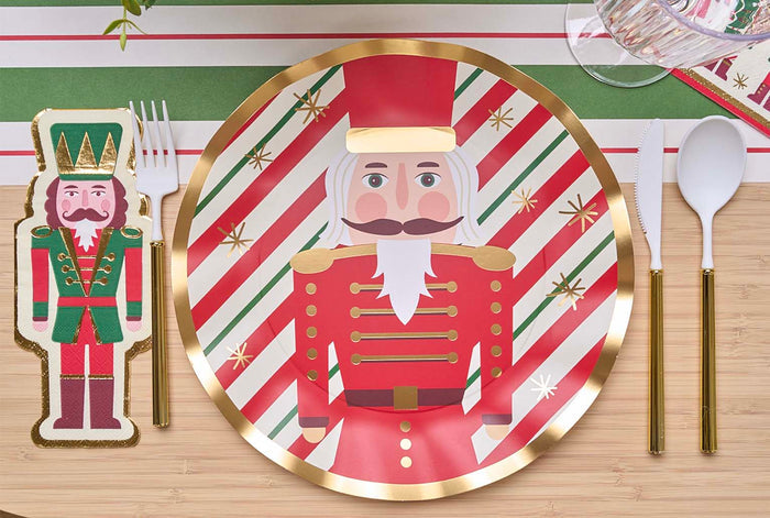 The Nutcracker Christmas Table Setting by Sophistiplate features designer disposable plates and napkins with a cartoon nutcracker, gold-trimmed utensils, and a red, white, and green striped runner for a festive holiday table.