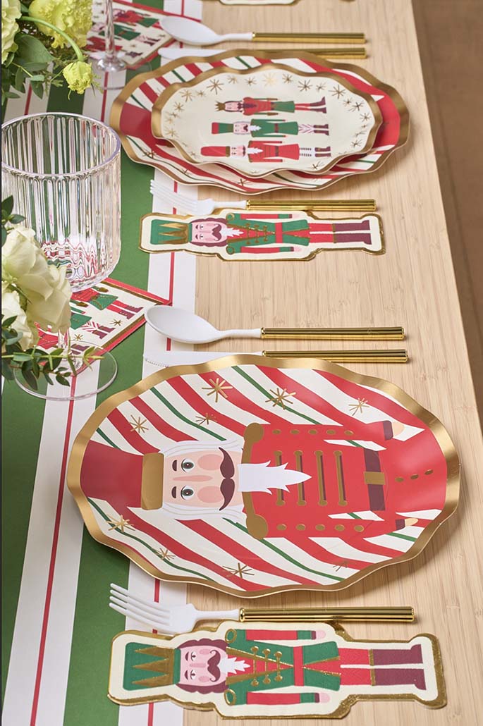 The Nutcracker Christmas Table Setting by Sophistiplate features festive designer plates and napkins with nutcracker figures, gold utensils, clear glassware, and a green-and-white striped runner with a floral centerpiece.