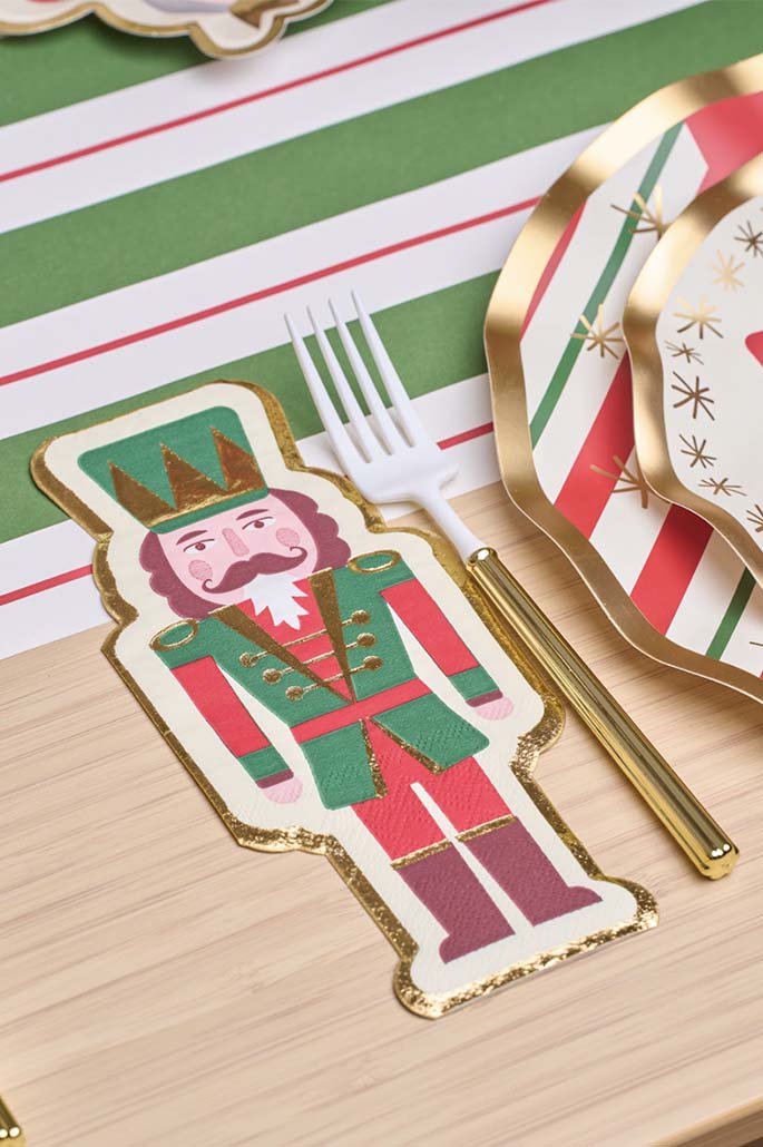 A festive table set with Sophistiplate's Guest Towel Nutcracker Christmas napkins (16/pkg), gold and white flatware, and red, green, and white striped plates atop a matching tablecloth.