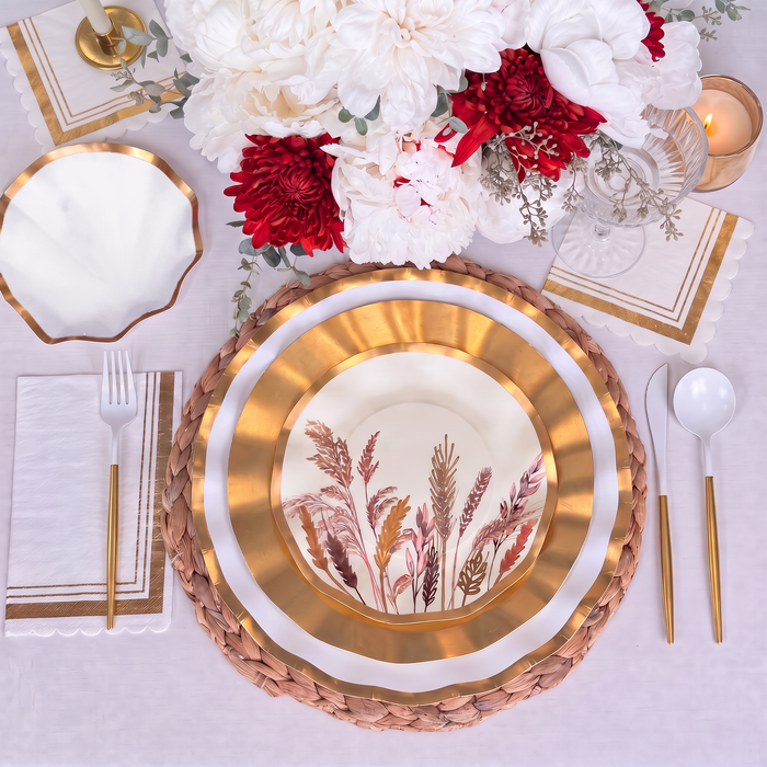 The Harvest Gold Table Setting by Sophistiplate features gold and white decorative botanical paper plates, gold flatware, a woven placemat, a white napkin, and a floral centerpiece with white and red blooms atop a crisp white tablecloth, creating an effortless tablescape in a box.