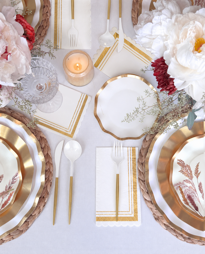 Create an exquisite tablescape with the Sophistiplate Harvest Gold Table Setting, featuring gold-trimmed decorative paper plates, gold and white cutlery, floral napkins, a glowing candle, and large white and pink flowers artfully arranged on a pristine white tablecloth—all elegantly boxed for your convenience.