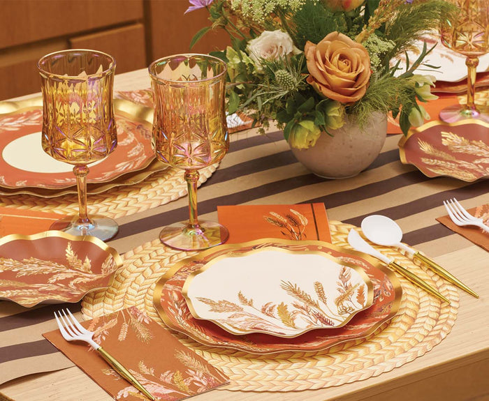 A fall-themed table setting featuring Sophistiplate’s Harvest Wheat Salad Plates with gold trim, paired with gold utensils, amber glasses, orange napkins, a floral centerpiece of roses, a woven placemat, and a brown striped table runner.