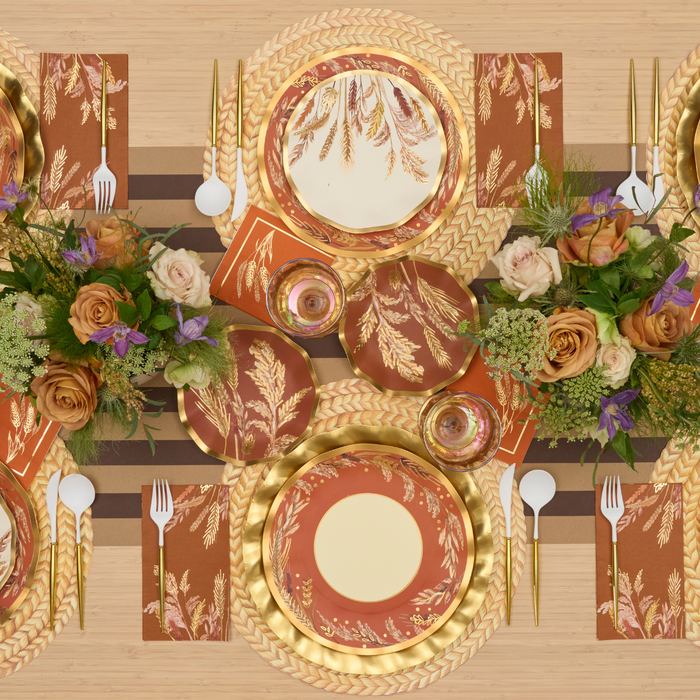 Elegant table setting with Sophistiplate Harvest Wheat Dinner Plates, featuring leaf patterns in gold and rust tones, paired with gold cutlery, floral centerpieces of roses and greenery, and rust napkins on a striped runner atop a wooden table.