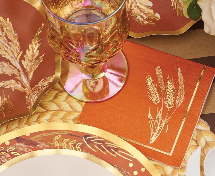 A close-up of the Sophistiplate Harvest Wheat Table Setting highlights designer disposable plates, an amber glass, and an orange napkin with a gold wheat motif—perfect for a warm, autumnal look.