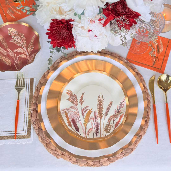 The Harvest Wheat Table Setting by Sophistiplate features botanical designer plates, a woven placemat, orange-handled cutlery, gold-rimmed glass, orange napkin, and floral accents on a white tablecloth.
