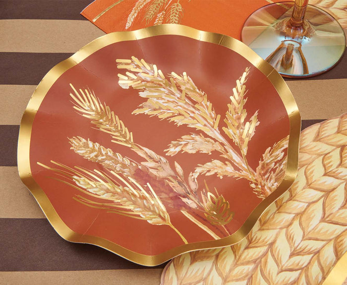 The Sophistiplate Harvest Wheat Table Setting features a decorative plate with gold and beige wheat on a burnt orange background. Matching designer disposable plates and a wine glass are also partially visible.
