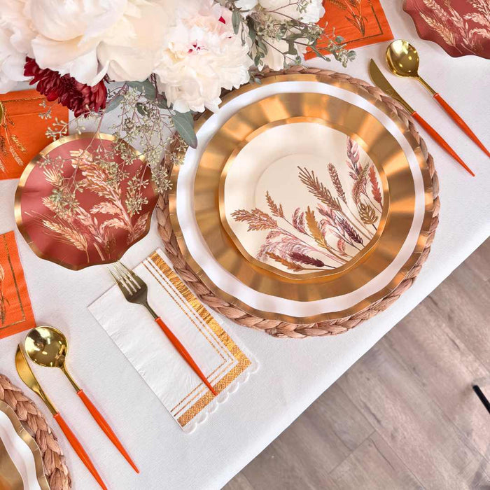 Create a festive table with the Sophistiplate Harvest Wheat Table Setting: gold and rust designer disposable plates, orange-handled cutlery, woven placemat, white napkin with orange trim, and a floral centerpiece on a white tablecloth.