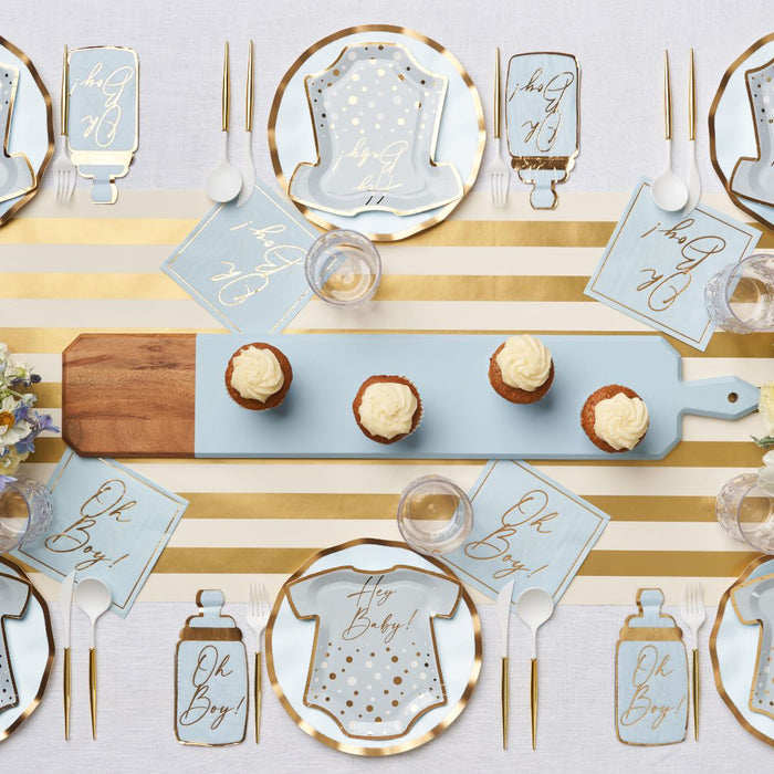 The Hey Baby Blue Table Setting by Sophistiplate LLC features designer disposable onesie-shaped plates in blue and gold, "Oh Boy!" napkins, gold utensils, cupcakes with frosting, and a gold-striped table runner for your baby shower.