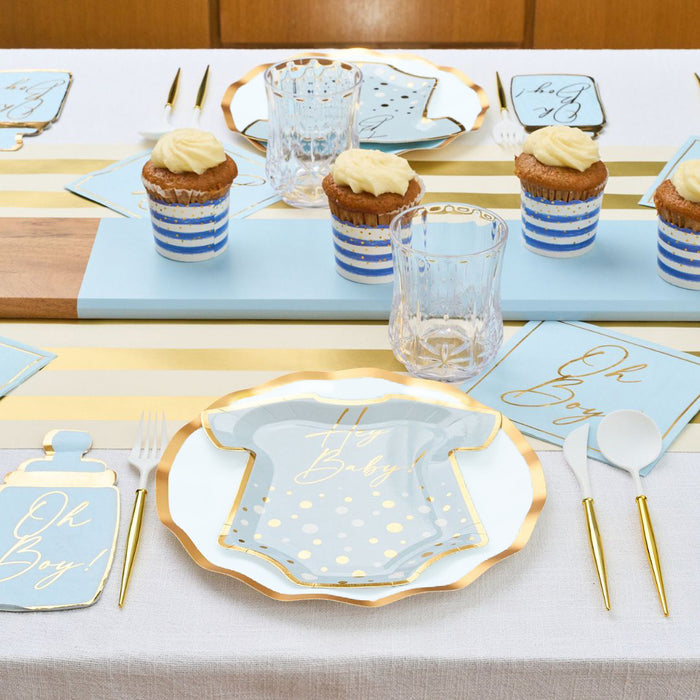 The Hey Baby Blue Table Setting by Sophistiplate LLC includes blue, white, and gold decorations with party-in-a-box supplies—onesie-shaped plates, striped cupcake liners, “Oh Boy!” napkins, and gold utensils atop a white tablecloth.