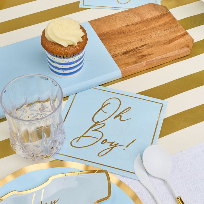 The Hey Baby Blue Table Setting by Sophistiplate LLC features a gold and white striped tablecloth, wooden board, cupcake in a blue-striped wrapper, designer disposable plates, white cutlery, clear glass, and an "Oh Boy!" blue napkin.
