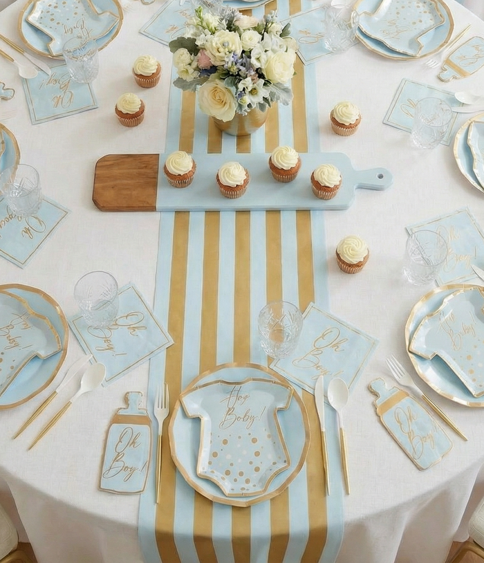 The Sophistiplate Hey Baby Blue Table Setting creates a chic baby shower with blue and gold designer plates, bottle-shaped cutlery holders, cupcakes, flowers, and a striped runner for an all-in-one stylish party setup.