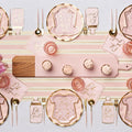 The Hey Baby Blush Table Setting by Sophistiplate LLC includes a striped table runner, onesie-shaped plates, "Oh Girl" napkins, pink cups, gold cutlery, cupcakes, and a wooden serving board for easy party styling.