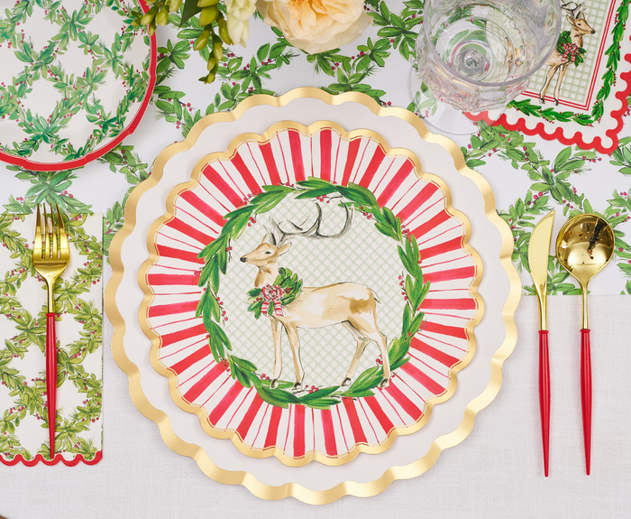 A festive table is set with gold and red cutlery, a floral napkin, and Sophistiplate's Holly Berry Dinner Plates (8pk), featuring a reindeer and holiday wreath. Greenery accents and a glass of water complete the cheerful look.